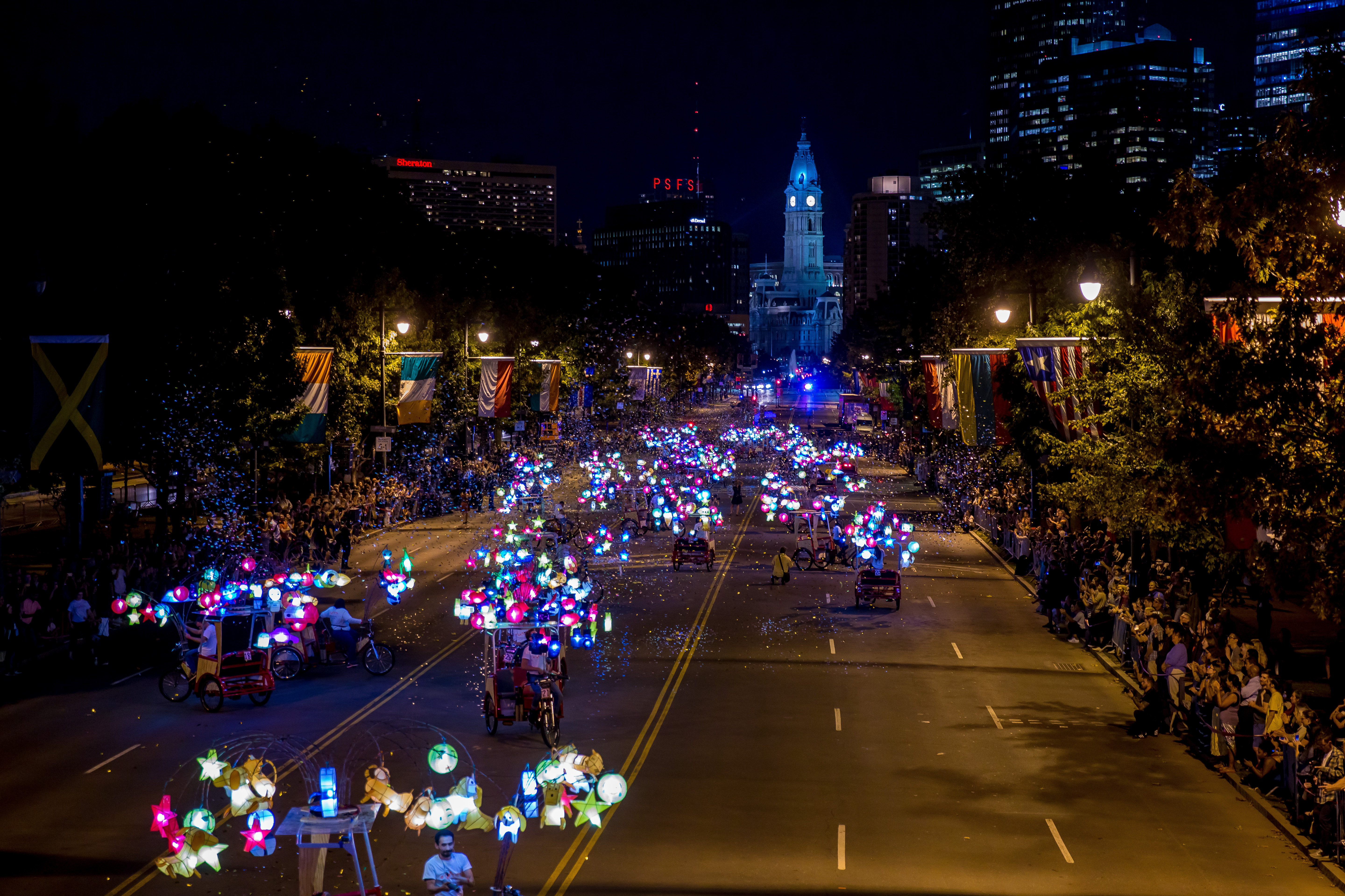 27 customized pedal vehicles adorned with over 900 handcrafted Chinese lanterns riding down Benjamin Franklin Parkway for a moving public art experience. Crowds of people line the Parkway with the pedal bikes driving down the middle.