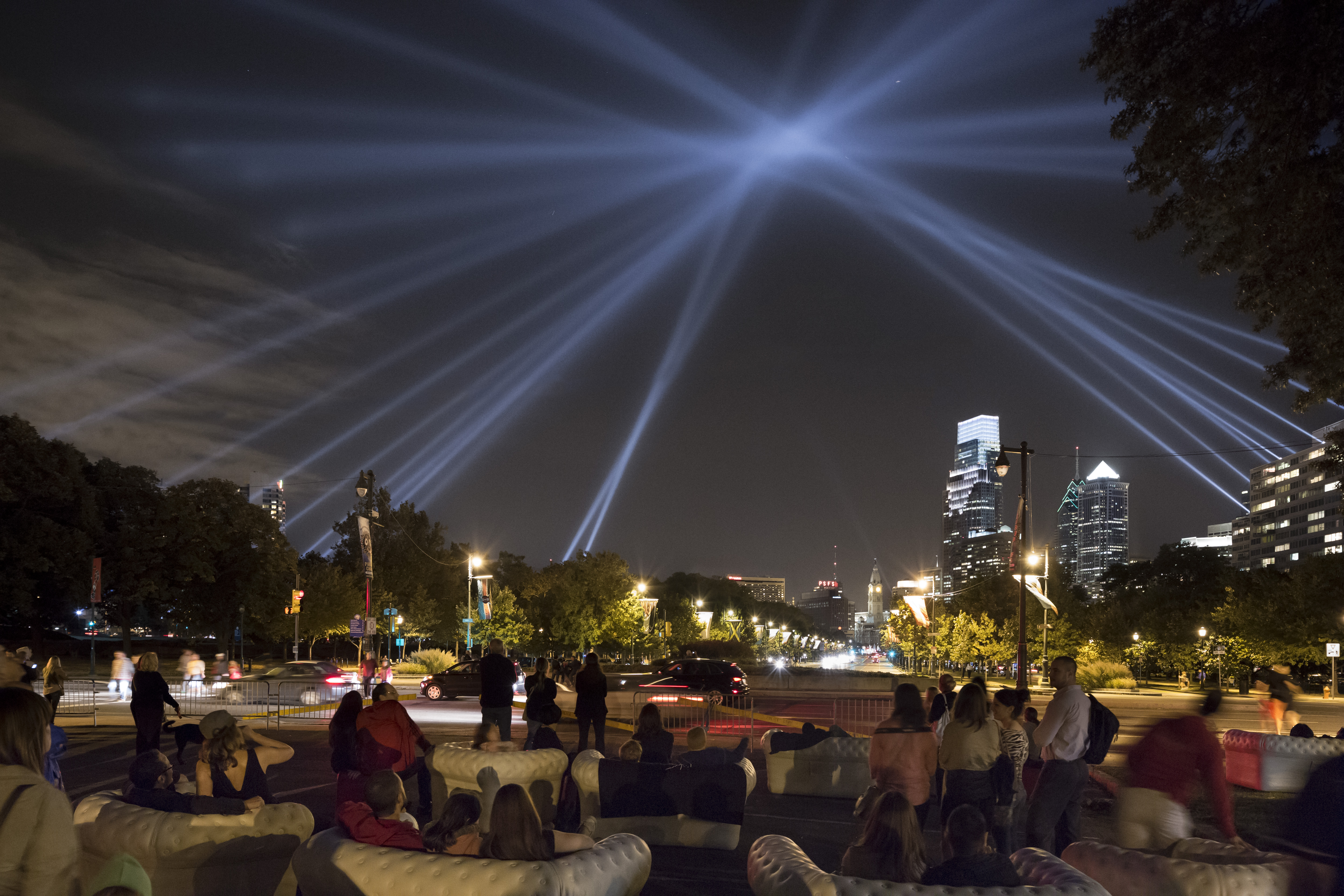 Large group of people sitting on furniture in from of the Philadelphia Museum of Art looking up at a light display in the sky