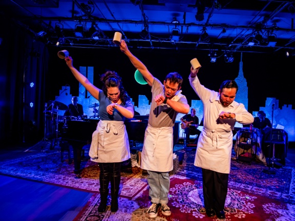 Two men and a woman on stage looking at their wrist watches with mugs in the air