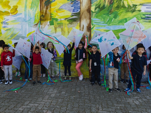 Small children in front of yellow and green mural with arts and craft kites raised above their heads