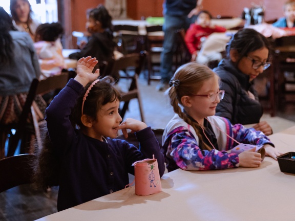 Two elementary school aged girls sit down and raise their hand during teaching artist instruction