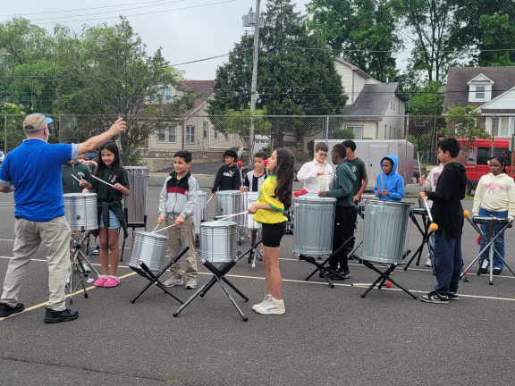 Large group of elementary school students on a basketball black top playing various drum and percussion instruments while being instructed by a man in a blue shirt.