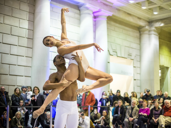 Male ballet dancer in white tights lifting up a woman ballet dancer with her arms in a right degree angle in front of a crowd in large space with white columns.