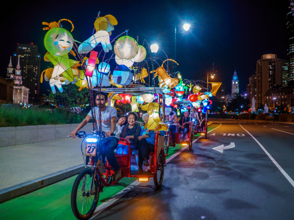 Man driving three wheeled bike covered in handcrafted Chinese lanterns driving down the Benjamin Franklin Parkway with three Asian women in the back seat smiling and enjoying the ride. 