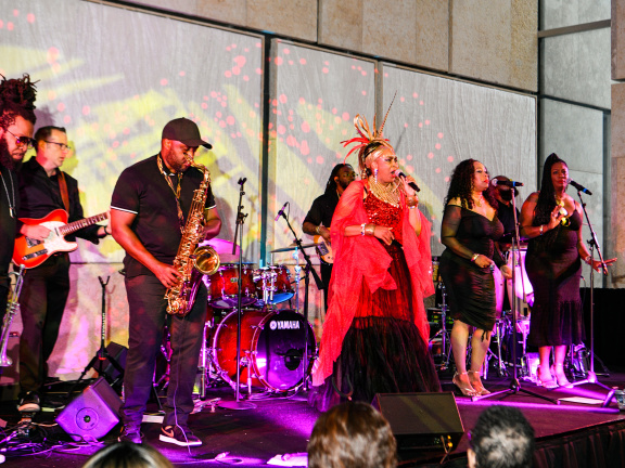 Black woman in a red dress and feathered headwear singing on stage with two black women as background singers dressed in black to her right. To her left is a black man in a black hat playing a brass instrument, and behind her is a drum set. 