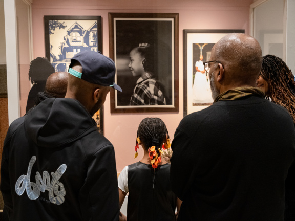 Two Black men and a young Black girl admire an art display case featuring photography including a portrait of another young Black girl. 