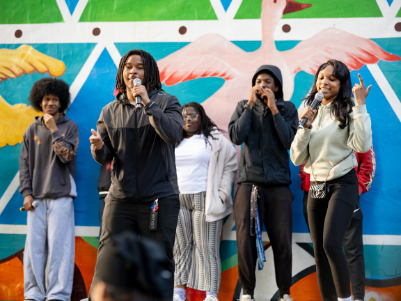 Three Black teenage boys and one Black teenage girl with microphones performing on a stage with a colorful mural in the background..