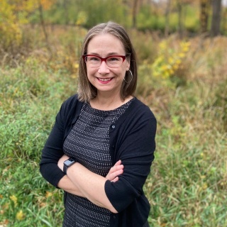 Staff headshot of Meghan Beals in a black cardigan and charcoal dress with her arms crossed in a green space.