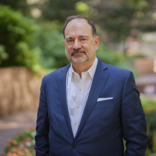 Middle aged white man in a blue suit jacket with a white button up with greenery in the background. 