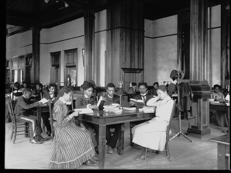 Interior view of library reading room with male and female students sitting at tables, reading, at the Tuskegee Institute. 