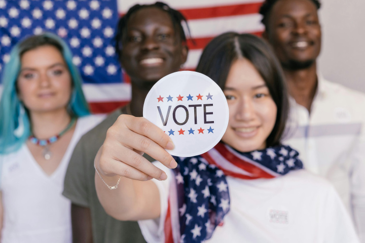 A person holding out a sticker that says "vote" with three other people and an American Flag in the background