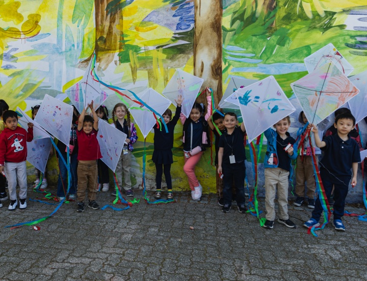 Small children in front of yellow and green mural with arts and craft kites raised above their heads