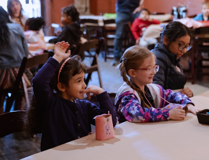 Two elementary school aged girls sit down and raise their hand during teaching artist instruction