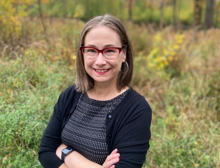 Staff headshot of Meghan Beals in a black cardigan and charcoal dress with her arms crossed in a green space.