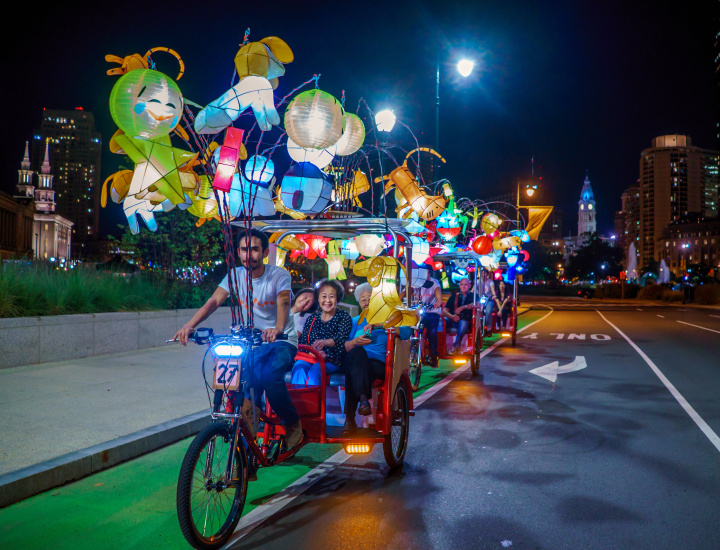 Man driving three wheeled bike covered in handcrafted Chinese lanterns driving down the Benjamin Franklin Parkway with three Asian women in the back seat smiling and enjoying the ride. 