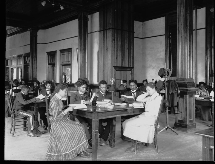 Interior view of library reading room with male and female students sitting at tables, reading, at the Tuskegee Institute. 