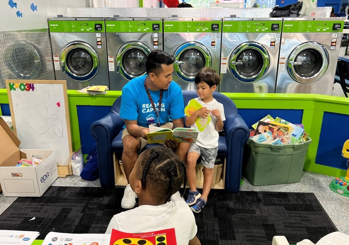 An adult and a child read a book together while sitting in a children's nook inside a laundromat. 